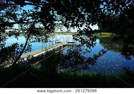 Long wooden bridge in lake with calm water view from behind trees, Sweden, Scandinavia, Europe. Peaceful outdoor image of Malaren lake in Vastmanland