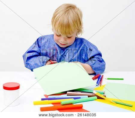 Menino feliz cortando seu caminho em uma folha de papel em uma mesa cheia de lápis coloridos, glu