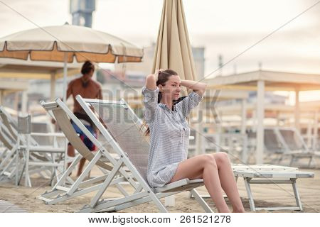Romantic young beautiful girl with long fair hair in short dress sitting in chaise-longue on the beach. Attractive sleepy calm woman dreaming at dawn on the shore in Italy, Europe. Soft focus.