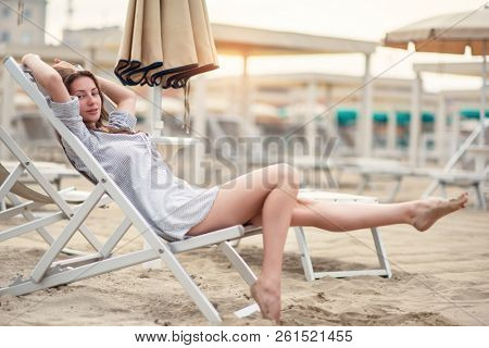Romantic young beautiful girl with long fair hair in short dress sitting in chaise-longue on the beach. Attractive sleepy calm woman dreaming at dawn on the shore in Italy, Europe. Soft focus