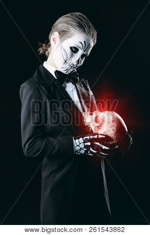 Portrait of a child boy with carnival make-up of a skeleton wearing elegant tail-coat. Studio shot over black background. Halloween. Dia de los muertos. The Day of the Dead.
