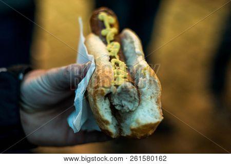Person Hand Holding Bratwurst Or Rostbratwurst A Traditional German Sausage, With Mustard On A Bun O