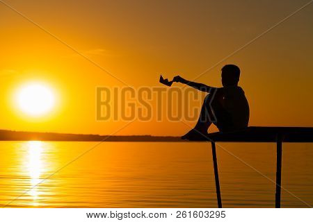 A Little Boy Sits On A Bridge And Holds Paper Ship In His Hand At Sunset At The River. A Kid Plays W
