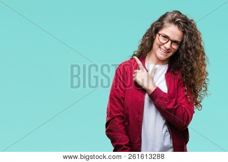 Beautiful brunette curly hair young girl wearing jacket and glasses over isolated background cheerful with a smile of face pointing with hand and finger up to the side with happy and natural expressio