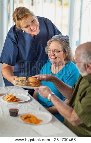 Female Doctor or Nurse Serving Senior Adult Couple Sandwiches at Table.