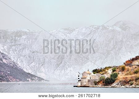 A Small Black And White Striped Lighthouse Near The Old Stone Chapel On The Background Of The Water 
