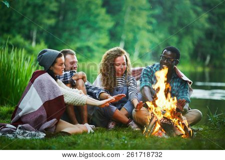 Several young backpackers sitting by campfire by waterside and warming their hands