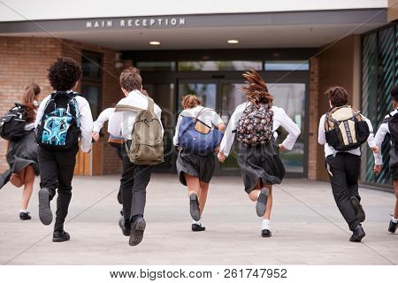 Group Of High School Students Wearing Uniform Running Into School Building At Beginning Of Class