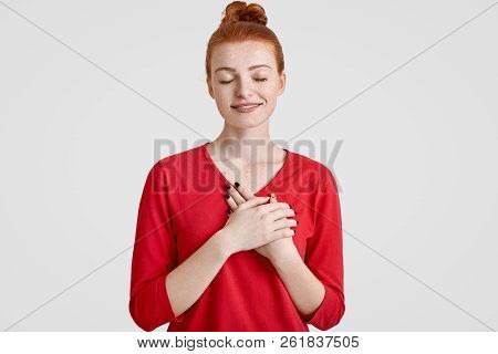 Studio Shot Of Calm Careless Woman With Ginger Hair Bun, Keeps Both Palms On Chest, Closes Eyes, Dre