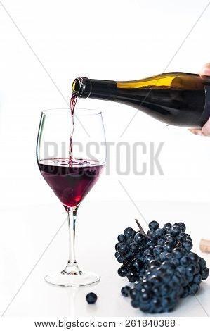 Red Wine Poured By A Person From A Bottle Into A Glass With A Cluster Of Black Grapes On White Table