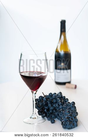Glass Of Red Wine With A Bottle And A Cluster Of Black Grapes On White Table. Selecive Focus.