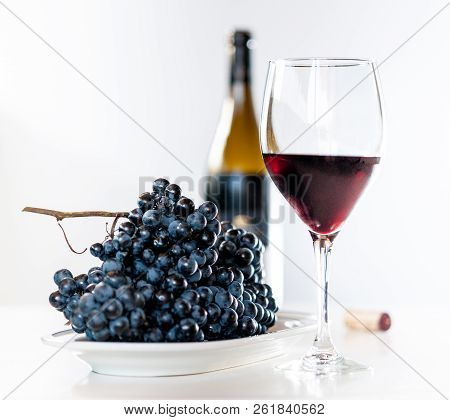 Glass Of Red Wine With A Bottle And A Cluster Of Black Grapes On White Table. Selecive Focus.