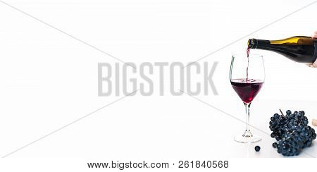 Red Wine Poured By A Person From A Bottle Into A Glass With A Cluster Of Black Grapes On White Table