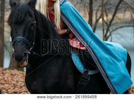 A Girl With Long Hair, Blonde Hair, Sits On The Back Of A Black Horse, In A Blue Cape And A Vintage 
