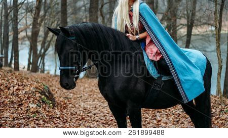 A Girl With Long Hair, Blonde Hair, Sits On The Back Of A Black Horse, In A Blue Cape And A Vintage 