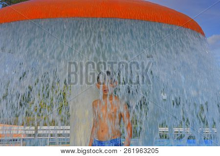 Happy Little Boy Enjoying Summer Day In The Swimming Pool. Boy Standing Under Sprinkler In Spray Poo