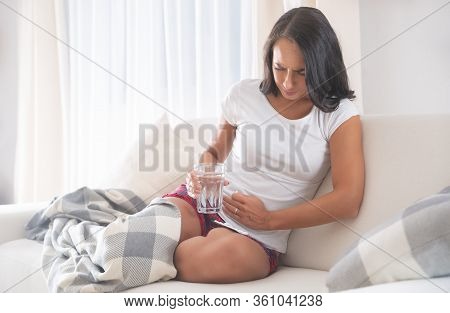 Woman Frowning In Ibd Pain Partially Covered By A Blanket Sitting On A Couch, With One Hand Covering