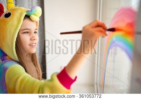 Stay Home. Teen Girl In Kigurumi Unicorn Draws The Rainbow On The Window. Flashmob Chasetherainbow.