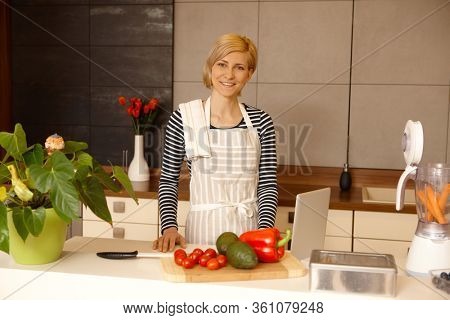 Smiling young woman standing in kitchen preparing healthy food, using tablet, looking at camera.