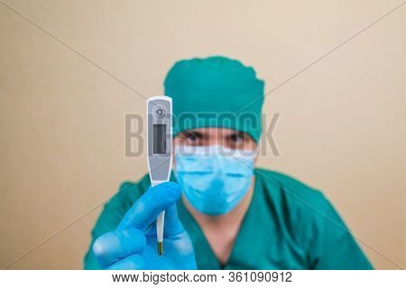 Doctor In Green Suit And Blue Mask Gloves With Thermometer In Hand On Yellow Background. Selective F