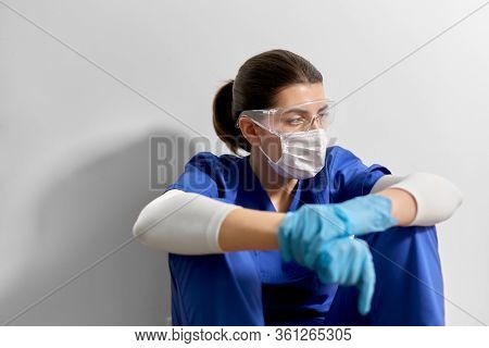medicine, healthcare and pandemic concept - sad young female doctor or nurse wearing goggles and face protective mask for protection from virus disease sitting on floor at hospital