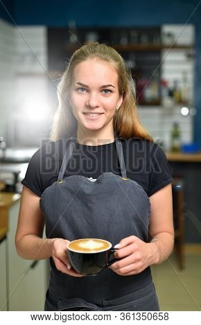A Young Barista Girl Holds A Large Mug Of Fresh Coffee And Smiles. Ssmall Business Concept And Young