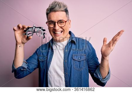Young modern optical optomestrist man holding optometrical glasses over pink background very happy and excited, winner expression celebrating victory screaming with big smile and raised hands