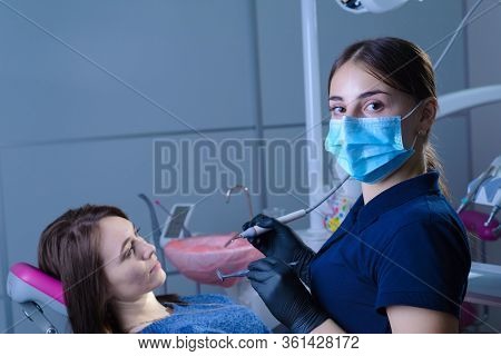Portrait Of A Dentist, In A Medical Mask, Holds A Dental Tool In His Hand, The Patient Sits In A Cha