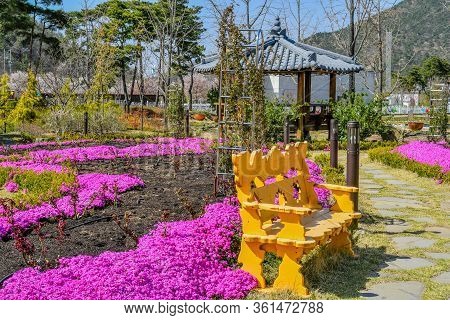 Yellow Park Bench Next To Stone Walkway Through Flower Garden With Gazebo In Background.