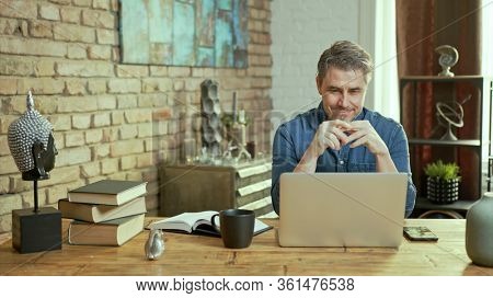 Older man tele working with laptop computer at home office in living room, thinking, looking busy.