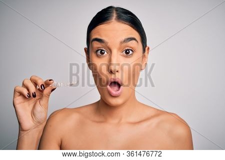 Young beautiful brunette woman holding dental aligner orthodontic to teeth corretion scared in shock with a surprise face, afraid and excited with fear expression
