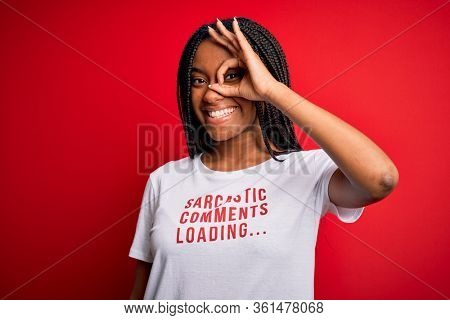 Young african american woman wearing sarcasm coments text on t-shirt over red background with happy face smiling doing ok sign with hand on eye looking through fingers