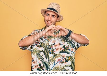 Young handsome man with blue eyes on vacation wearing summer florar shirt and hat smiling in love showing heart symbol and shape with hands. Romantic concept.
