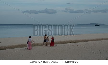 Women In Sarongs With Man, Girl And Child Walking Along Beach With Boat In The Background