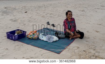 Woman Sitting Cross Legged On Beach Mat And Smiling With Purple Basket And Purse With Beach Shoes In