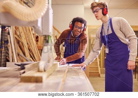 Tutor With Male Carpentry Student In Workshop Studying For Apprenticeship At College Using Bench Saw