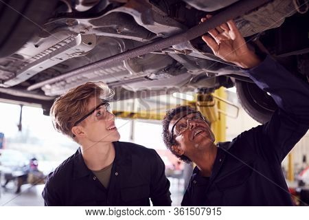 Male Tutor With Student Looking Underneath Car On Hydraulic Ramp On Auto Mechanic Course