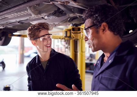 Male Tutor With Student Looking Underneath Car On Hydraulic Ramp On Auto Mechanic Course