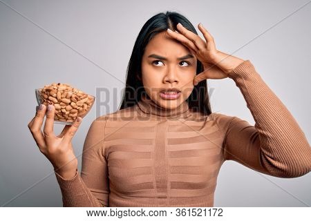 Young asian girl holding bowl of healthy peanuts eating as snack for diet over white background stressed with hand on head, shocked with shame and surprise face, angry and frustrated. Fear and upset