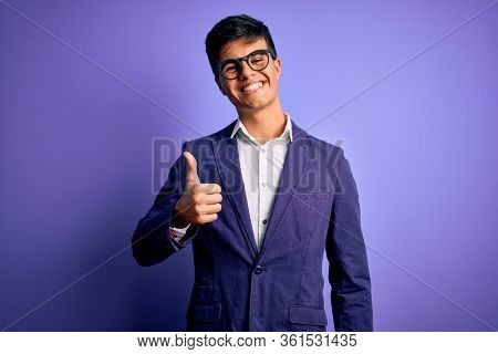 Young handsome business man wearing jacket and glasses over isolated purple background doing happy thumbs up gesture with hand. Approving expression looking at the camera showing success.