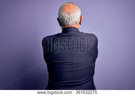 Grey haired senior business man wearing glasses and elegant suit and tie over purple background Hugging oneself happy and positive from backwards. Self love and self care