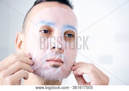 closeup of a handsome caucasian man applying or removing a bio-cellulose sheet mask to his own face