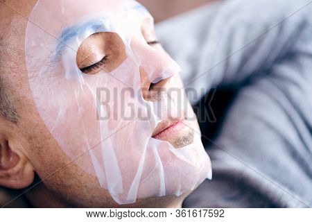 closeup of a handsome caucasian man wearing a bio-cellulose sheet mask while is relaxing