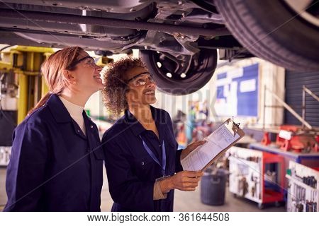 Female Tutor With Student Looking Underneath Car On Hydraulic Ramp On Auto Mechanic Course