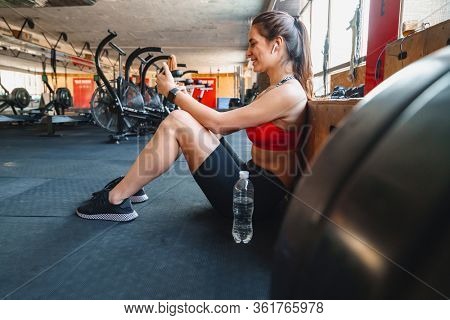 Portrait of cheerful cute sportswoman using wireless earphone and cellphone while sitting in athletic hall