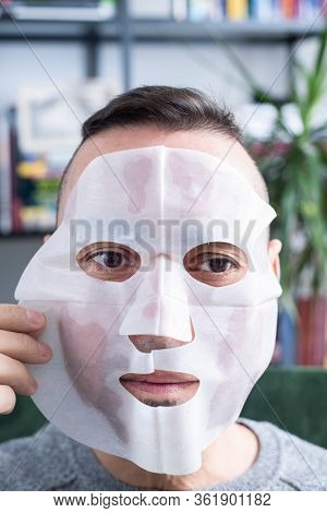 closeup of a handsome caucasian man applying a bio-cellulose sheet mask to his own face