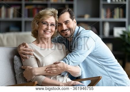 Adult Son Hugs Elderly Mother While She Resting On Armchair
