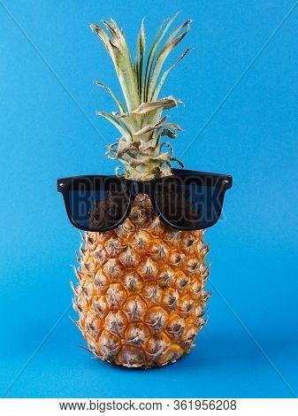 Front View Of A Ripe Orange Pineapple With Green Leaves And Black Sunglas On It, On A Blue Backgroun