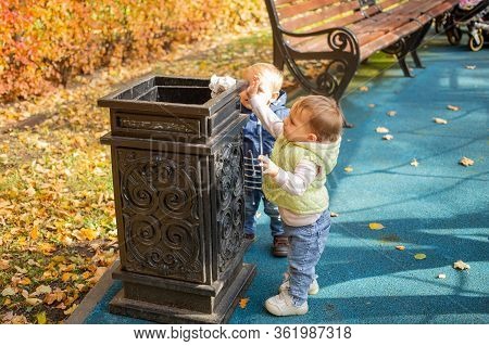 Two Little Cute Babies Are Standing Near The Black Street Container In The Park And Throwing Garbage