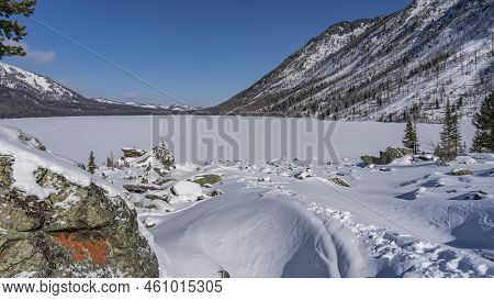 The Frozen Lake Surrounded By Mountains Is Covered With Untouched White Snow. In The Foreground Are 